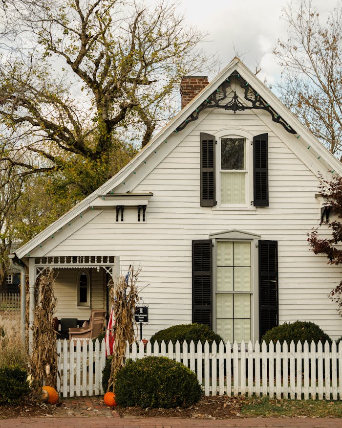 Quaint white cottage featuring pumpkins, white picket fence, and rustic charm.